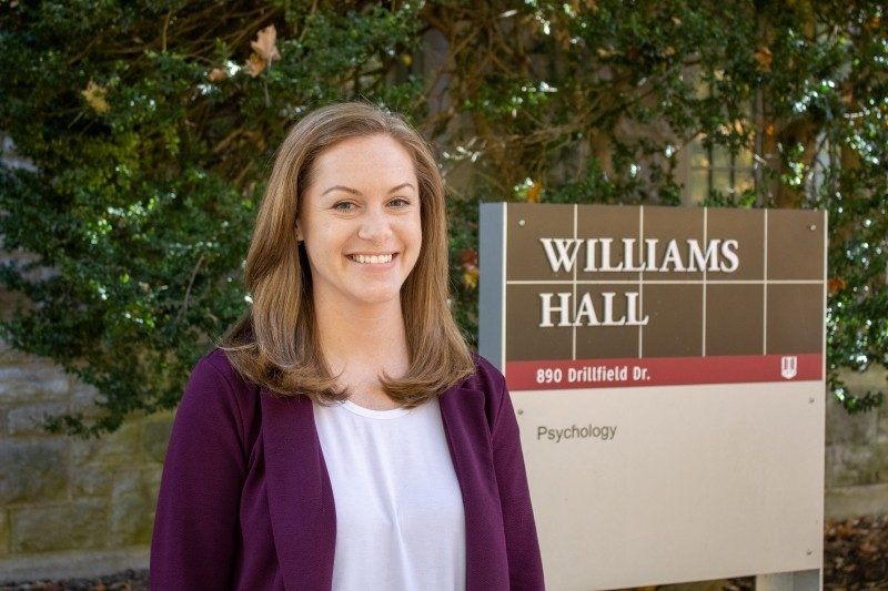 Meagan Brem stands outside in front of the Williams Hall building sign on Virginia Tech's campus wearing a white shirt and purple blazer jacket.