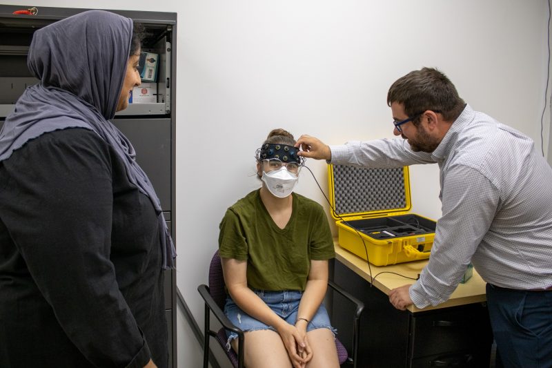 Ben Katz Lab Photo with Graduate Students Marrium Mansoor, left, observes Ben Katz, right, as he demonstrates how to use the fNIRS machine, which Diane Devine, center, is wearing on her head.